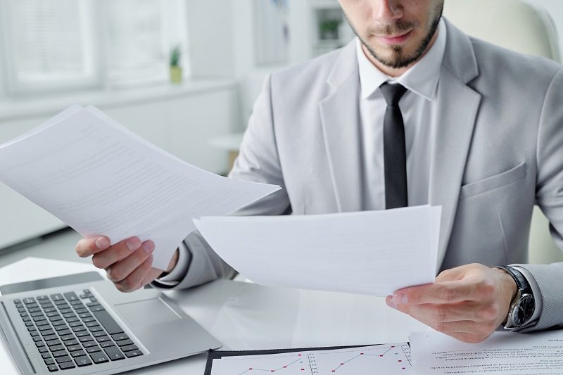 Close-up of concentrated young bearded manager sitting at desk in office and analyzing document content